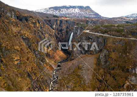 Magnificent Voringsfossen waterfall plunges into deep rugged canyon flanked by rocky cliffs. Winding road and sparse vegetation complete landscape 128735961