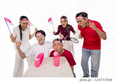 Portrait of an Indonesian southeast asian people celebrate Indonesia Independence Day 17th of August with game blowing ball on plastic glass isolated white background. Concept of Kemerdekaan Indonesia Portrait of an Indonesian southeast asian people celebrate Indonesia Independence Day 17th of August with game blowing ball on plastic glass isolated white background. Concept of Kemerdekaan Indonesia 128737098