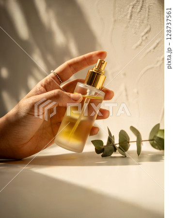 Elegant perfume bottle with golden spray held in hand next to eucalyptus leaves in warm light Elegant perfume bottle with golden spray held in hand next to eucalyptus leaves in warm light 128737561
