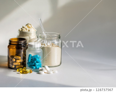 Assorted health supplements with capsules and protein powder in glass jars under soft light and shadow on white surface 128737567