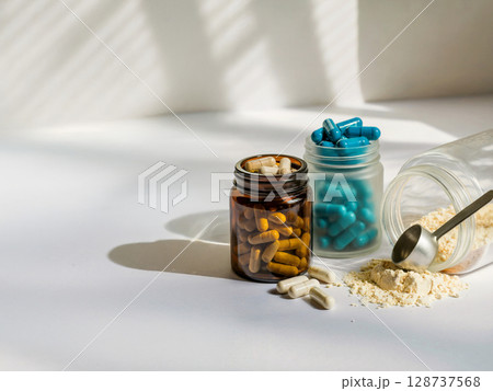 Glass jars with blue and golden capsules and protein powder spilling with scoop on white table and shadow light 128737568
