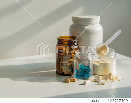 Assorted nutritional supplements with capsules and protein powder in various containers on white table under morning light 128737569