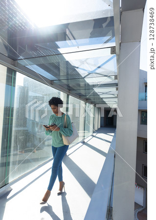 African American woman walking in glass corridor, checking smartphone and carrying tote, copy space African American woman walking in glass corridor, checking smartphone and carrying tote, copy space 128738469