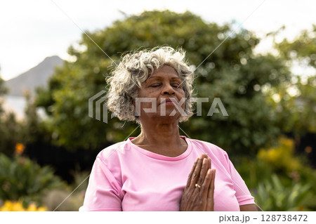 Senior woman meditating in garden park with hands clasped at chest, expressing calm focus 128738472
