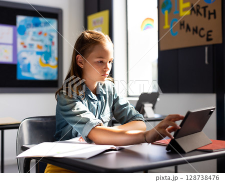 Child girl student tapping tablet on school desk in bright classroom, with open workbook Child girl student tapping tablet on school desk in bright classroom, with open workbook 128738476