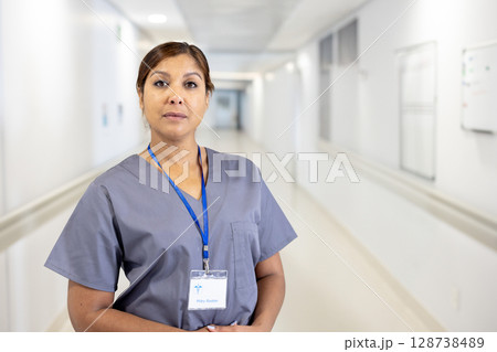 Senior woman standing in bright hospital corridor, wearing gray scrubs and blue lanyard badge Senior woman standing in bright hospital corridor, wearing gray scrubs and blue lanyard badge 128738489