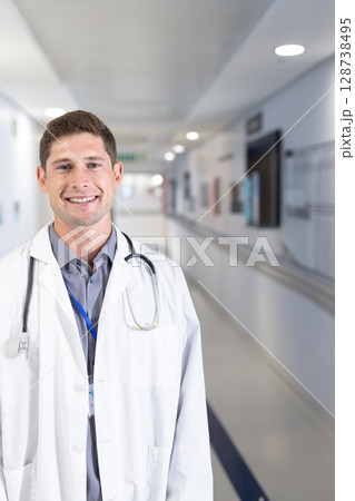 Male doctor standing and smiling in hospital corridor, with stethoscope and ID badge, copy space 128738495
