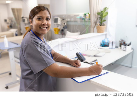 Mature adult nurse writing on clipboard with orange pen at hospital ward desk, with infusion pumps 128738502