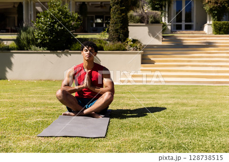 Young man meditating cross-legged on grey yoga mat in front yard, soaking up bright sunshine 128738515