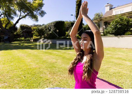 Woman meditating on purple yoga mat in backyard garden, holding hands pressed overhead, copy space 128738516