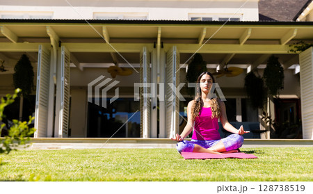 Woman meditating on pink yoga mat on backyard lawn in front of contemporary house, copy space 128738519