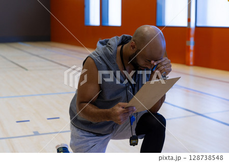 Mid-adult male coach kneeling on indoor basketball court, wiping sweat with clipboard and stopwatch Mid-adult male coach kneeling on indoor basketball court, wiping sweat with clipboard and stopwatch 128738548