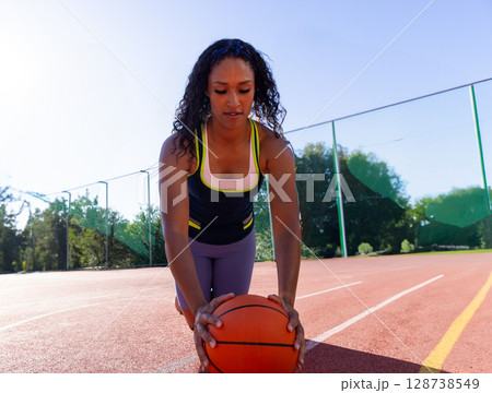 Young adult woman kneeling on outdoor basketball court, holding basketball beside chain?link fence Young adult woman kneeling on outdoor basketball court, holding basketball beside chain?link fence 128738549