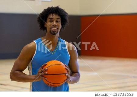 African American man standing on gym basketball court, holding basketball and smiling at camera African American man standing on gym basketball court, holding basketball and smiling at camera 128738552