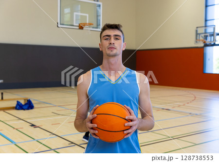 Young adult male standing in indoor court, holding orange basketball near hoop, wearing blue jersey Young adult male standing in indoor court, holding orange basketball near hoop, wearing blue jersey 128738553