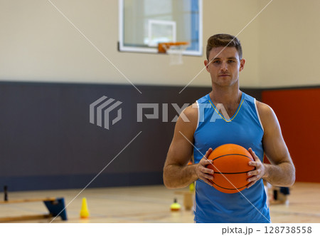 Male basketball player holding basketball on gym court, with training cones and hoop, copy space 128738558