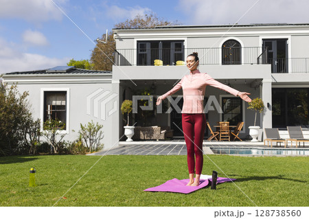Woman performing yoga stretch on pink mat on green lawn in suburban backyard, with water bottles 128738560