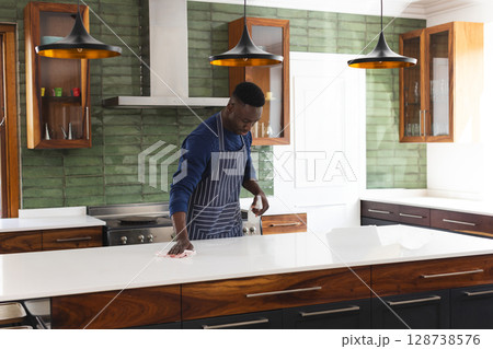 African American man cleaning quartz countertop in modern kitchen, with spray bottle and cloth 128738576