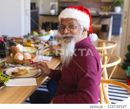 Senior man wearing Santa hat sitting at dining table at home, holding fork with red berries 128738587