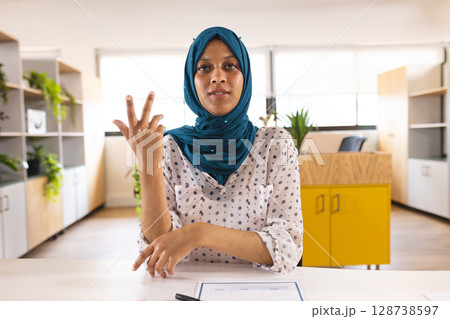 Woman wearing teal hijab showing three fingers at office desk with document pen and yellow cabinet 128738597