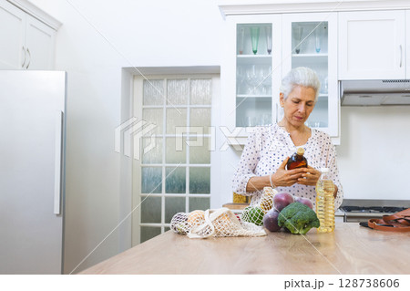 Senior woman reading oil bottle label at wooden island in modern white kitchen, with fresh produce 128738606