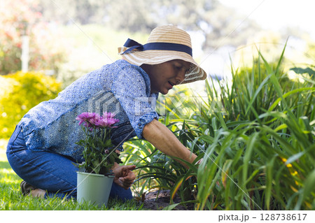 Senior woman kneeling on lawn in backyard garden, tending soil by green pot holding purple flowers 128738617
