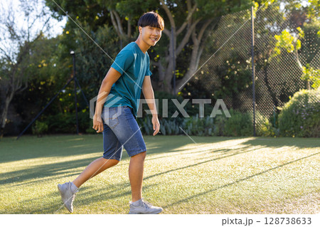 Young Asian man walking across synthetic turf in backyard, with chain-link fence, shrubs and trees 128738633
