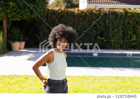 Young African American woman standing on lawn in private backyard, with swimming pool and hedgerow 128738640