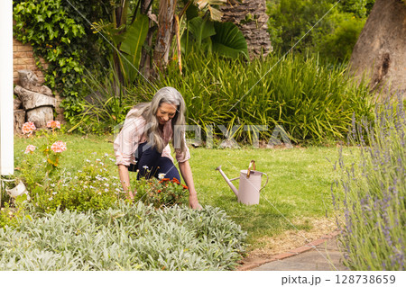 Senior woman kneeling and tending flower bed in private backyard garden, with pink watering can 128738659