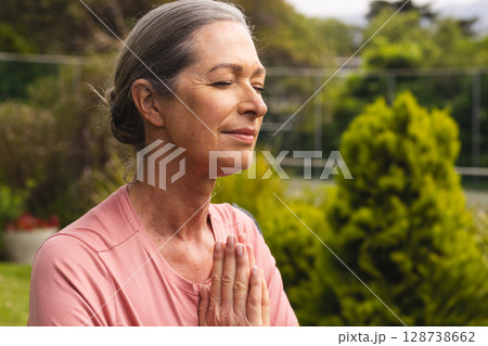 Senior woman meditating with hands clasped in backyard garden under sunlight, copy space Senior woman meditating with hands clasped in backyard garden under sunlight, copy space 128738662