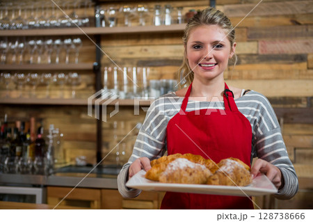 Young woman presenting freshly baked croissants behind cafe counter with wooden shelves, copy space Young woman presenting freshly baked croissants behind cafe counter with wooden shelves, copy space 128738666