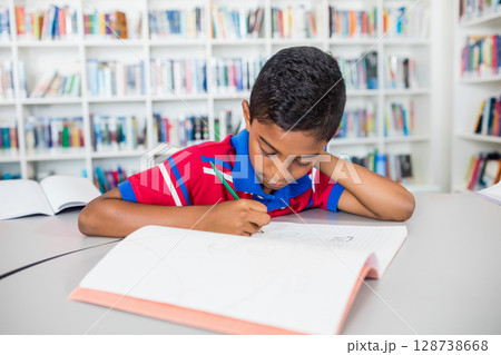 bi-racial boy writing in grid notebook at curved library table, using green pencil with open books 128738668