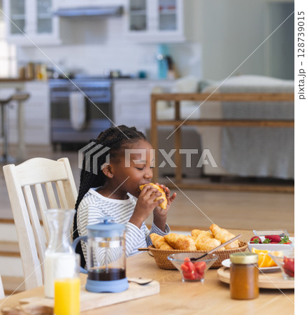 African American girl sitting at table in open kitchen eating croissant with bowl of strawberries African American girl sitting at table in open kitchen eating croissant with bowl of strawberries 128739015
