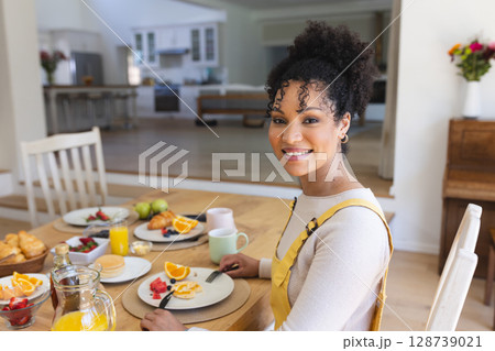 Young adult woman sitting at dining table in open-plan kitchen, smiling with fruit, copy space Young adult woman sitting at dining table in open-plan kitchen, smiling with fruit, copy space 128739021