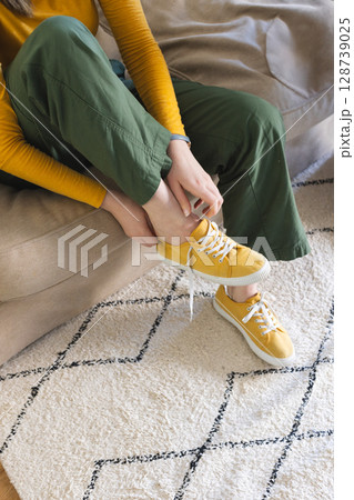 Mid-20s woman tying mustard-yellow canvas sneaker on beige couch in living room, with geometric rug Mid-20s woman tying mustard-yellow canvas sneaker on beige couch in living room, with geometric rug 128739025