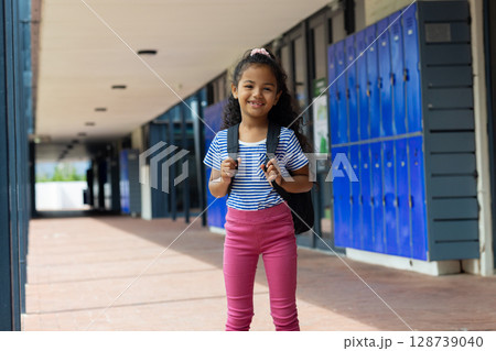 Child student standing in covered school corridor, with black backpack and joyful smile 128739040