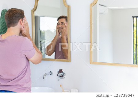 Man touching face at sink mirror in bright modern bathroom, with toothbrush and towel, copy space 128739047