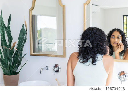 Asian Indian woman applying facial pad in home bathroom with wooden mirror and plant, copy space Asian Indian woman applying facial pad in home bathroom with wooden mirror and plant, copy space 128739048