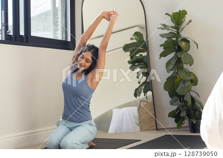 Indian woman in 20s kneeling stretching overhead on yoga mat near window in bedroom, copy space Indian woman in 20s kneeling stretching overhead on yoga mat near window in bedroom, copy space 128739050