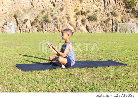 Young boy meditating on black yoga mat on sports field by rocky hill, with closed eyes 128739055