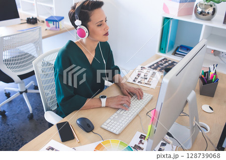 Woman typing on keyboard at desk with monitor in modern office, wearing headphones 128739068