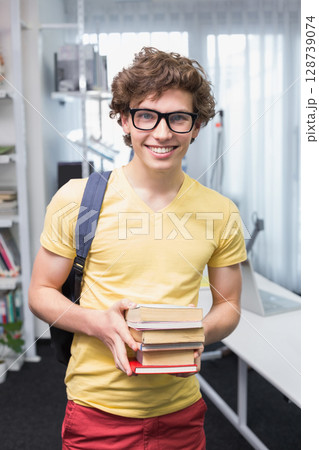 Man holding stack of hardcover books in study room, with backpack strap and open laptop visible 128739074