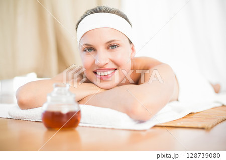 Young woman resting on massage table in bright spa, smiling beside amber oil in glass jar 128739080