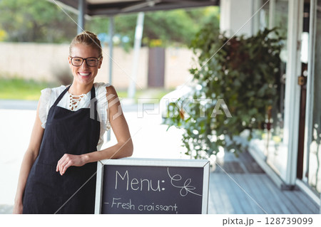 Woman smiling and leaning on chalkboard menu sign under cafe awning, wearing apron and glasses 128739099