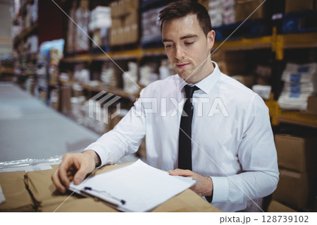 Man reviewing paperwork on clipboard in large warehouse, with pen, metal shelving and stacked boxes 128739102