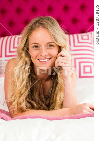 Woman lying on stomach in bedroom, smiling at camera with pink headboard and decorative pillows 128739135