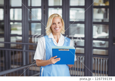 young adult woman standing in office hallway holding bright blue clipboard with both hands, smiling 128739139