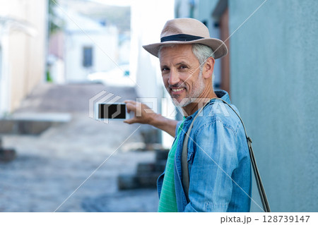 Senior man standing in sunlit stone-paved alley, holding smartphone and wearing fedora, copy space 128739147