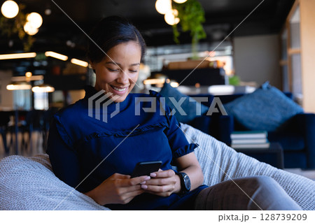 Woman sitting in cushioned lounge chair in co-working lounge, holding smartphone and smiling Woman sitting in cushioned lounge chair in co-working lounge, holding smartphone and smiling 128739209