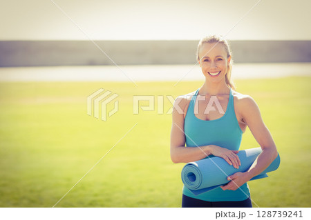 Mid-adult woman standing in sunny grassy field holding blue yoga mat, enjoying outdoor fitness Mid-adult woman standing in sunny grassy field holding blue yoga mat, enjoying outdoor fitness 128739241